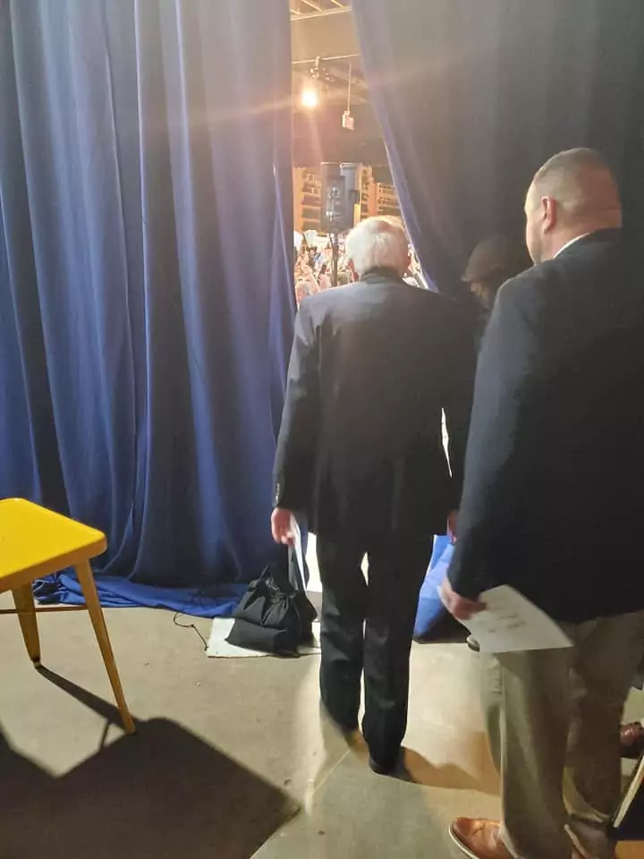 Two men in suits are walking through a room with blue curtains.